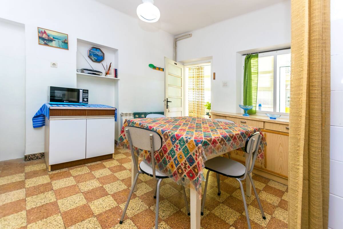 Een compacte keuken met een magnetron, een klein tafeltje met stoelen en een raam met gordijn.
