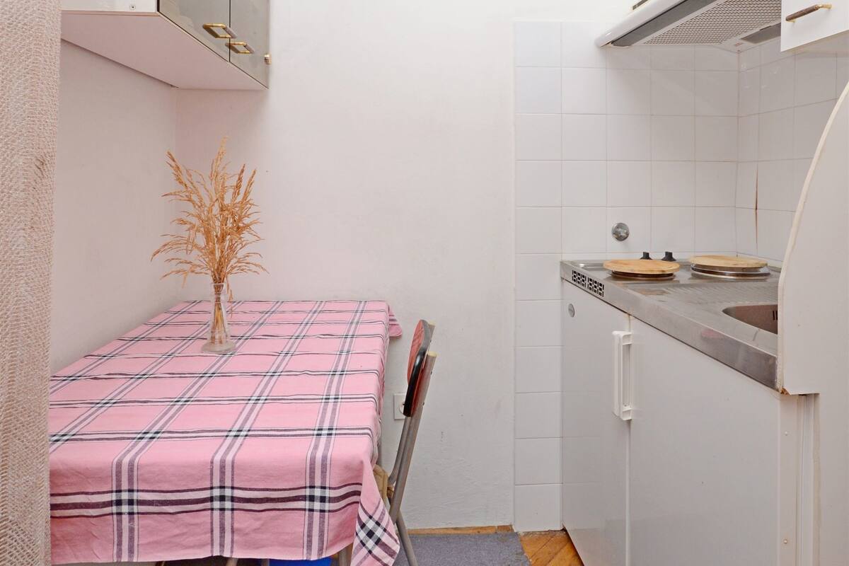 Una cocina pequeña con mantel a cuadros rosas, un taburete azul y un armario blanco con microondas.