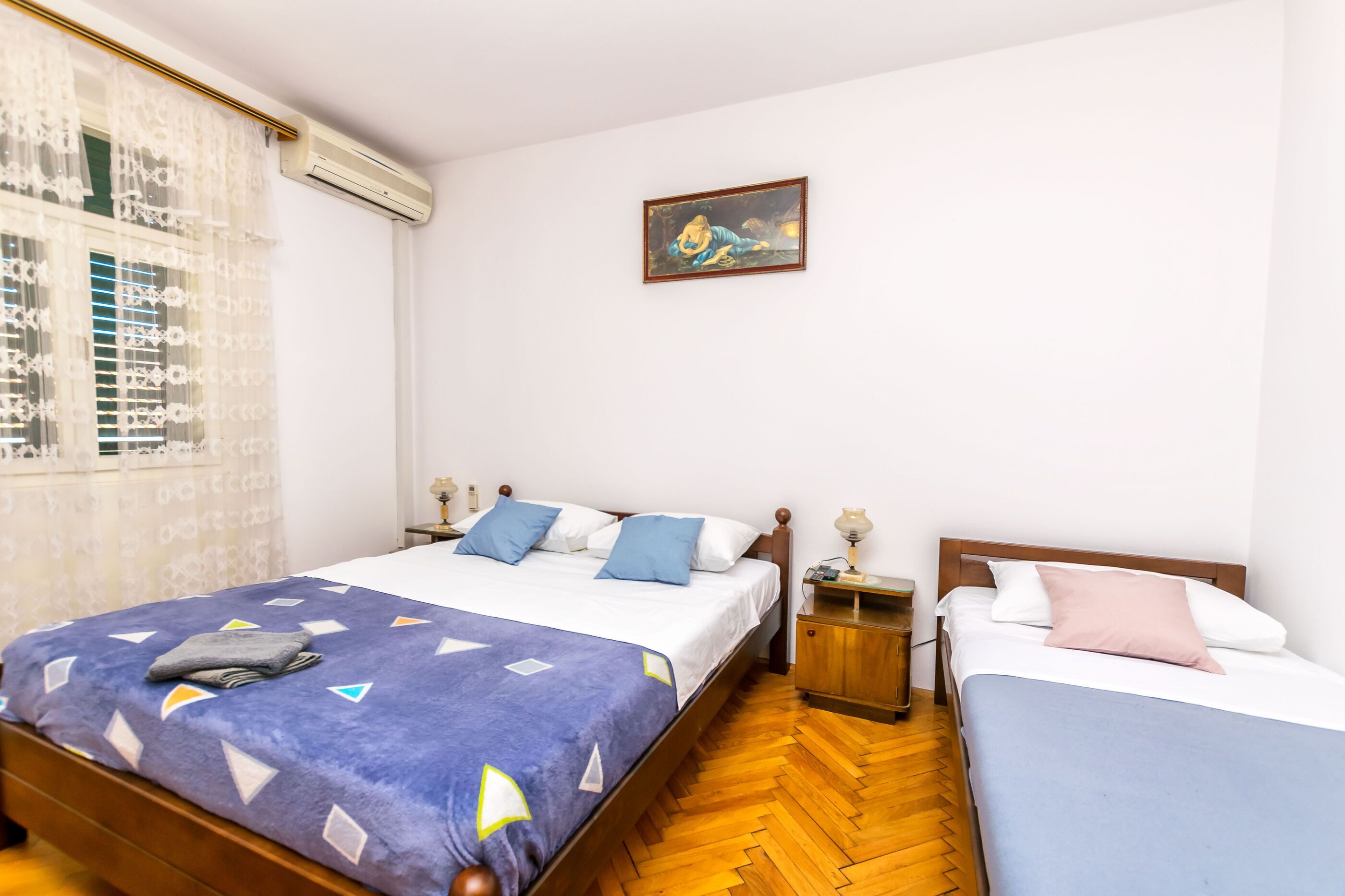 A bedroom with a bed, two bedside tables, a nightstand, a window with curtains, and a painting on the wall.