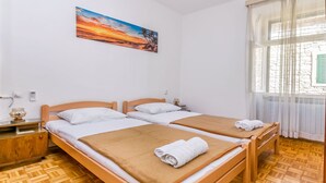 Une chambre avec deux lits en bois, du linge de lit blanc, un tableau représentant un coucher de soleil, une table de chevet en bois et une fenêtre ornée de rideaux légers.
