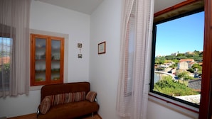 A room with a brown sofa, a wooden cabinet with glass doors, and a large window with a view of houses and greenery. - Apartments Loredana (Lumbarda)