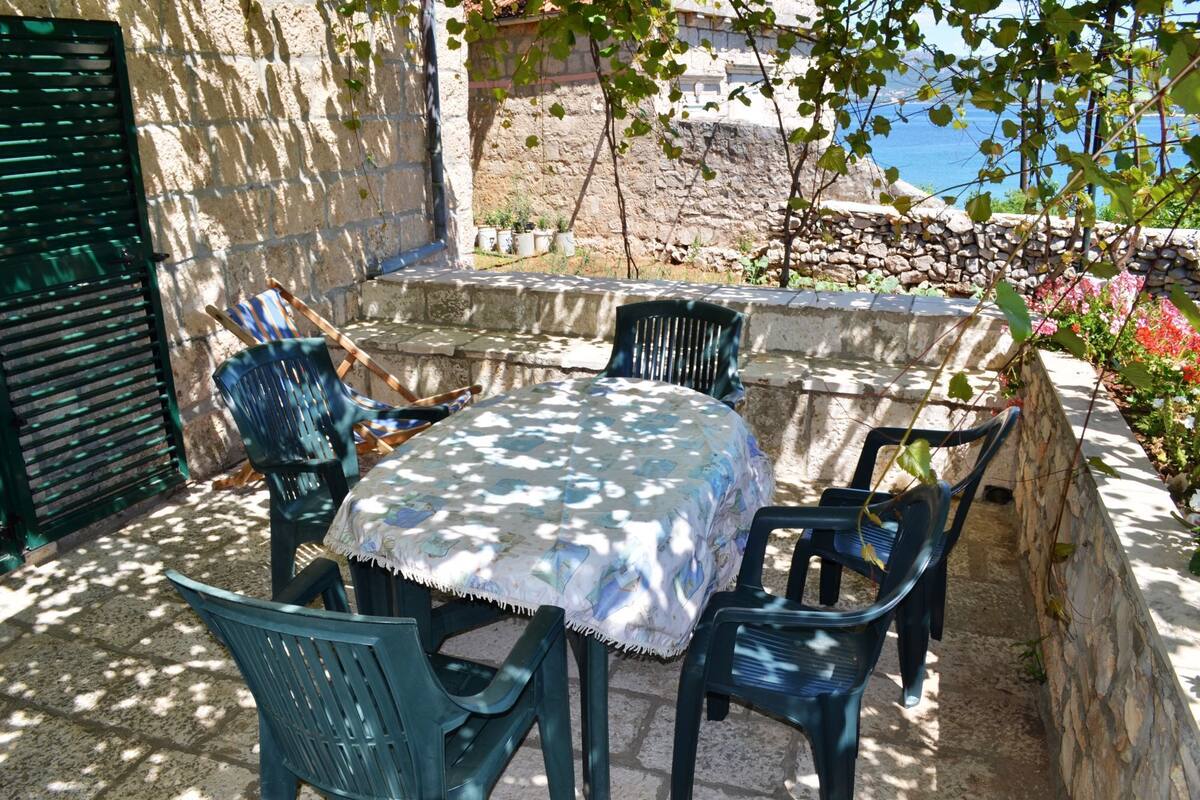 Un espace extérieur aménagé avec des sièges, une table recouverte d’une nappe à motifs, entourée de chaises en plastique vertes, le tout adossé à un mur de pierre avec vue sur la mer.