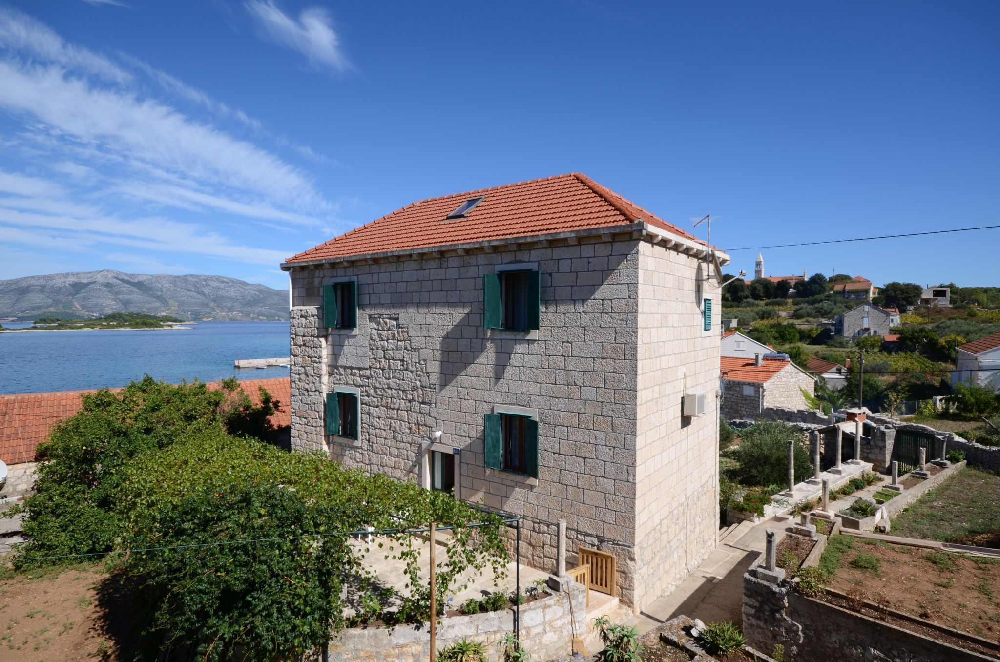 A stone building with a red-tiled roof, green shutters, and a view of a body of water and hills in the background.