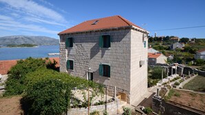 A stone building with a red-tiled roof, green shutters, and a view of a body of water and hills in the background. - Apartments Loredana (Lumbarda)