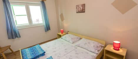 A bedroom with a bed, two bedside tables, a nightstand, a window with curtains, and a wall-mounted picture.