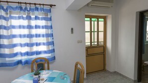 A dining area with a table set for two, a blue and white striped curtain, and a window with a wooden door. - Apartments Ksenija (Primosten)
