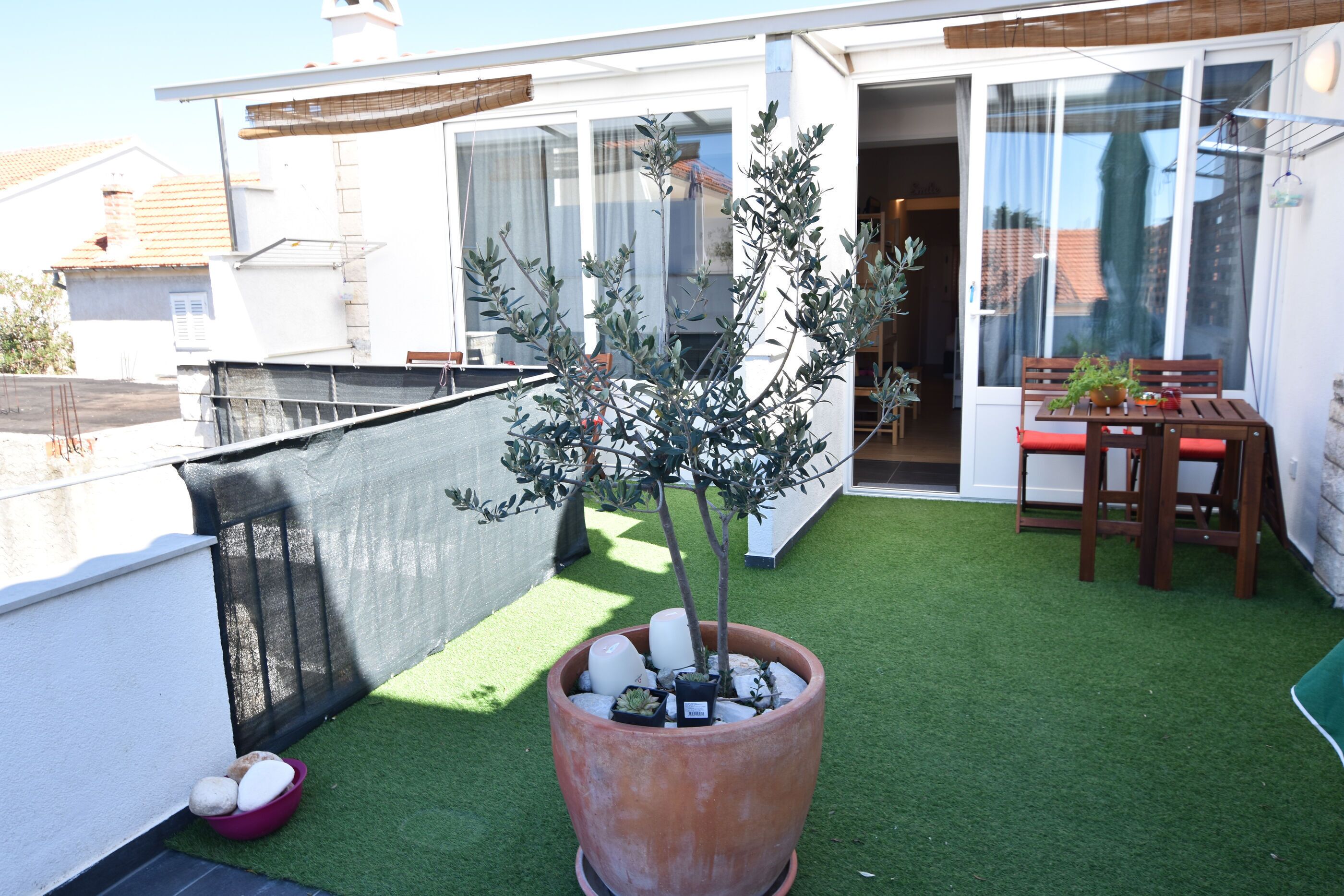 A rooftop terrace with artificial grass, a potted olive tree, a wooden table with chairs, and a sliding glass door leading to an indoor space.