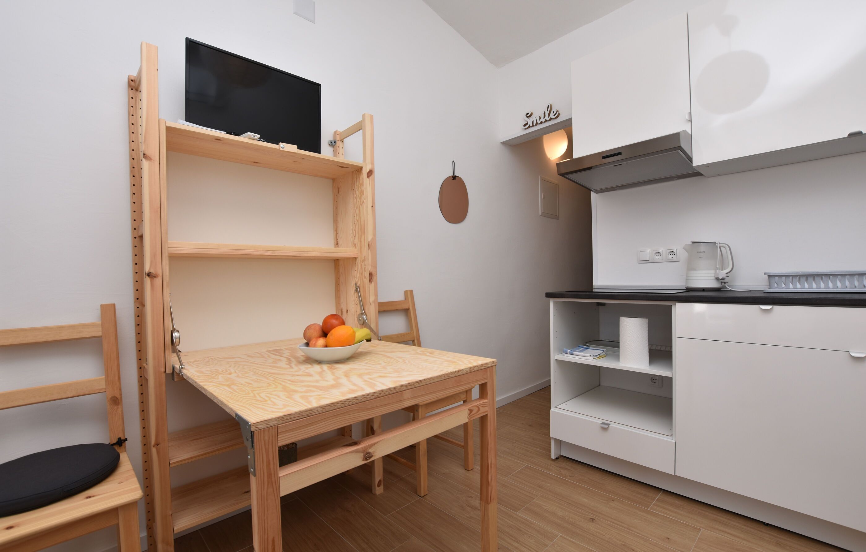 Cocina compacta con una mesa de madera, frutero, televisor y armarios blancos.