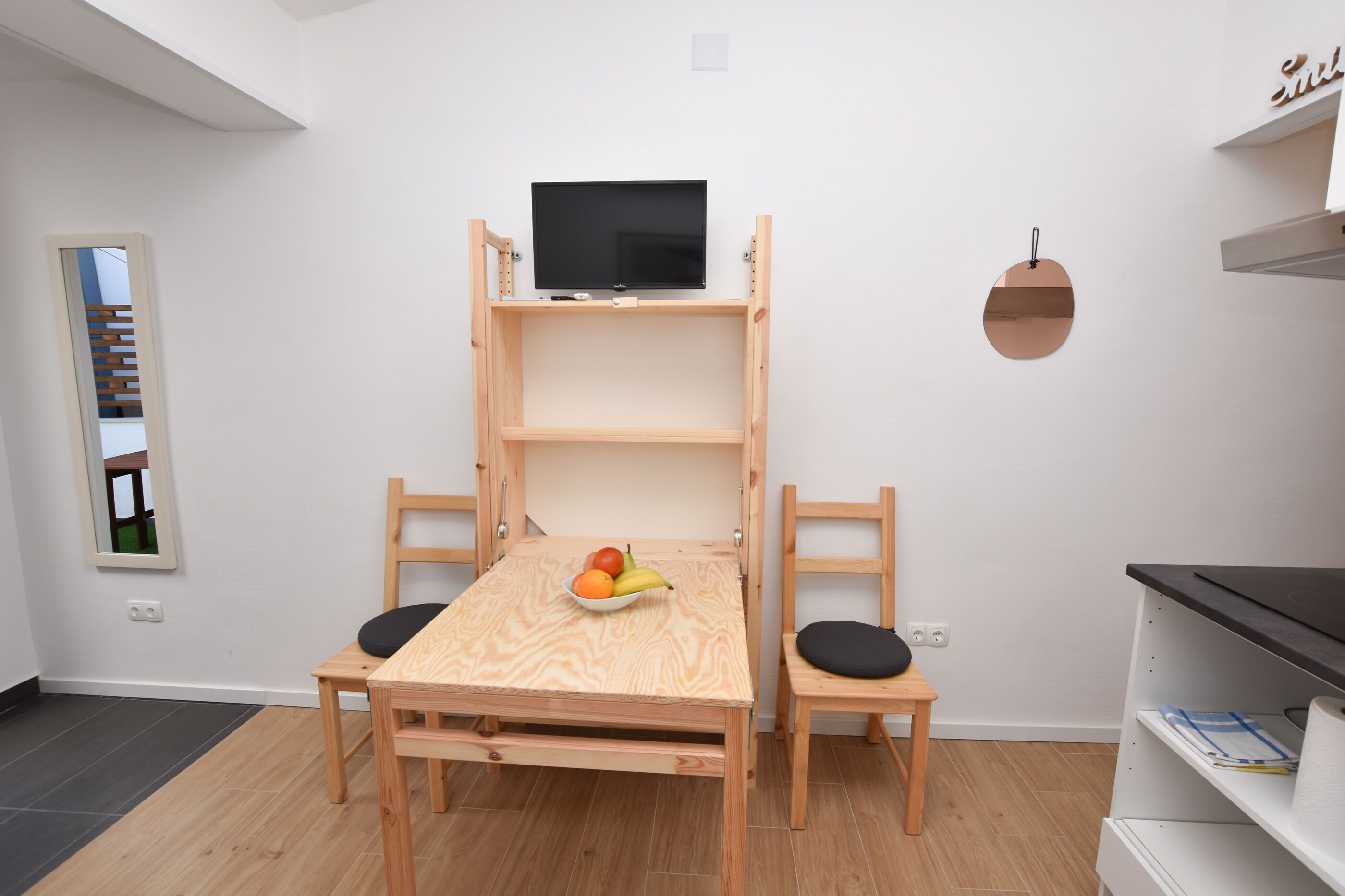 A compact room with a wooden bunk bed, a television on the top bunk, a small table with fruit, and a chair with a cushion.