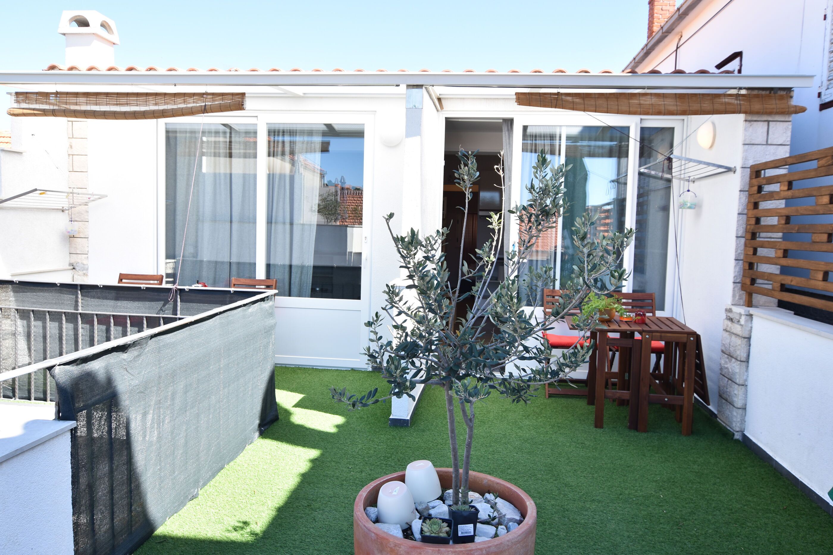 A small patio with artificial grass, a wooden table and chairs, a potted tree, and a sliding glass door leading to a room.