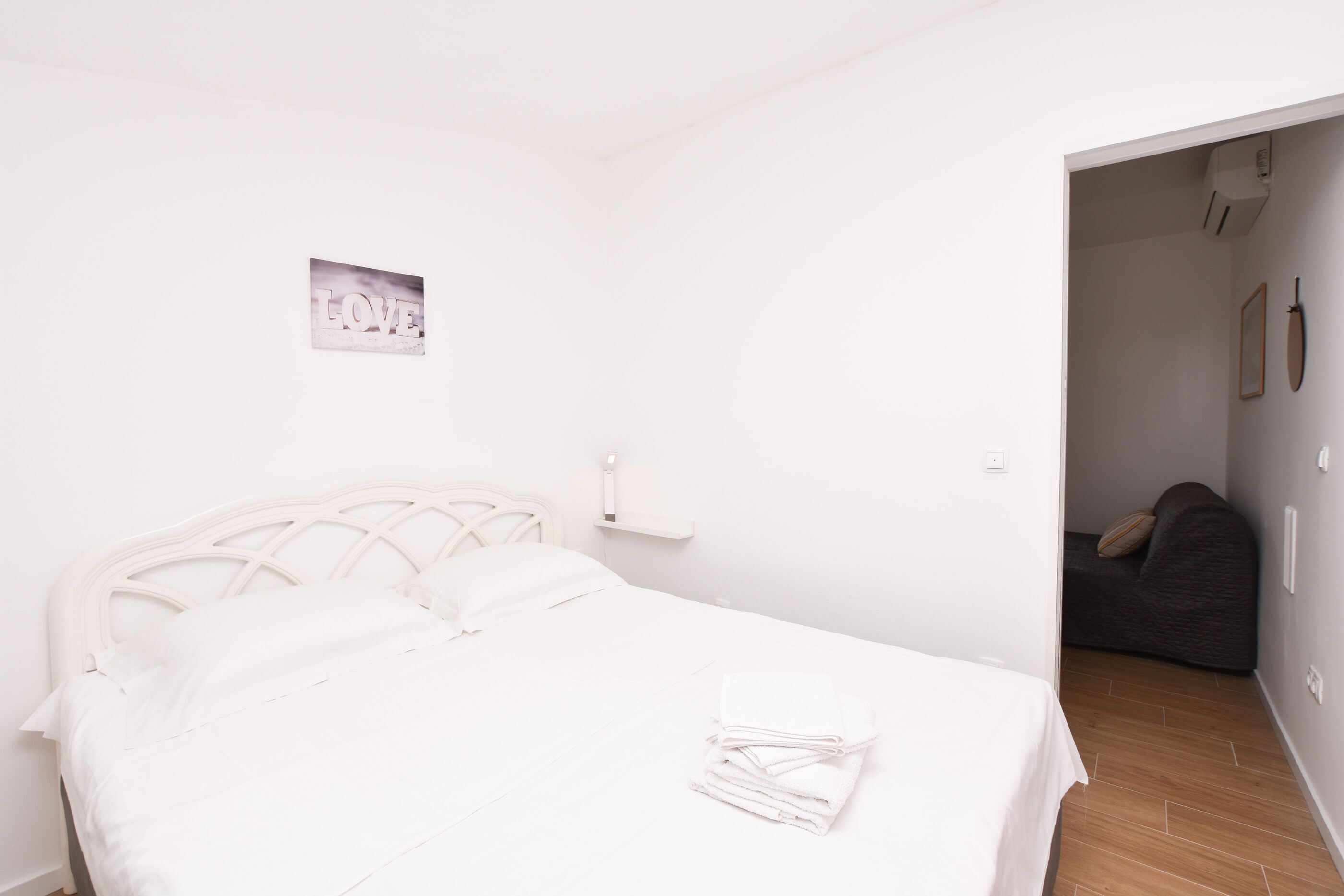 A white bed with a headboard, a small bedside table, and a folded white towel on the bed.