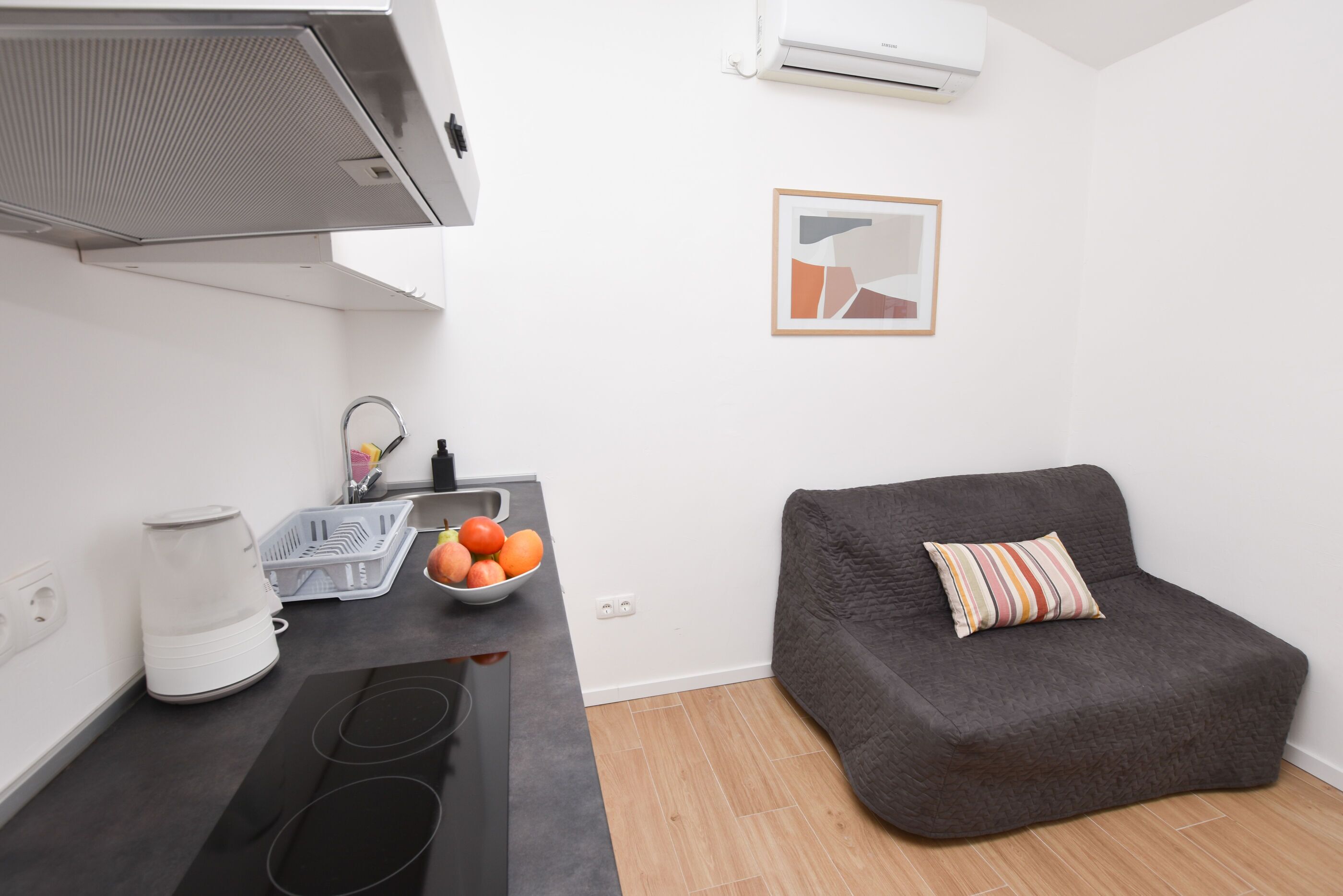 Une cuisine de taille réduite avec un plan de travail noir, un canapé gris agrémenté d’un coussin à rayures et une œuvre d’art abstraite encadrée accrochée au mur.
