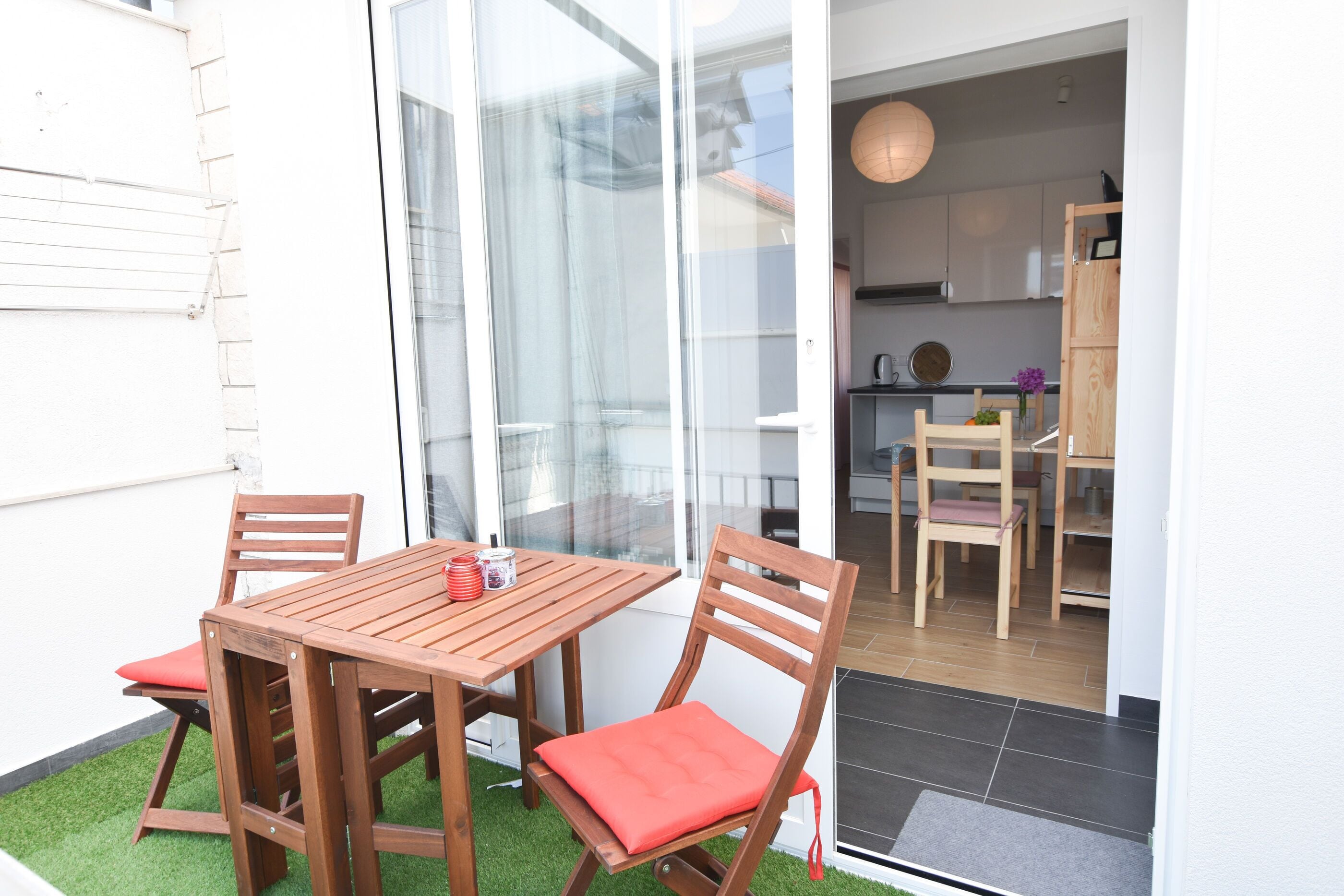 Una mesa y sillas de madera con cojines rojos en un balcón con césped artificial, una puerta de cristal que da a una cocina con armarios de madera y una lámpara colgante.