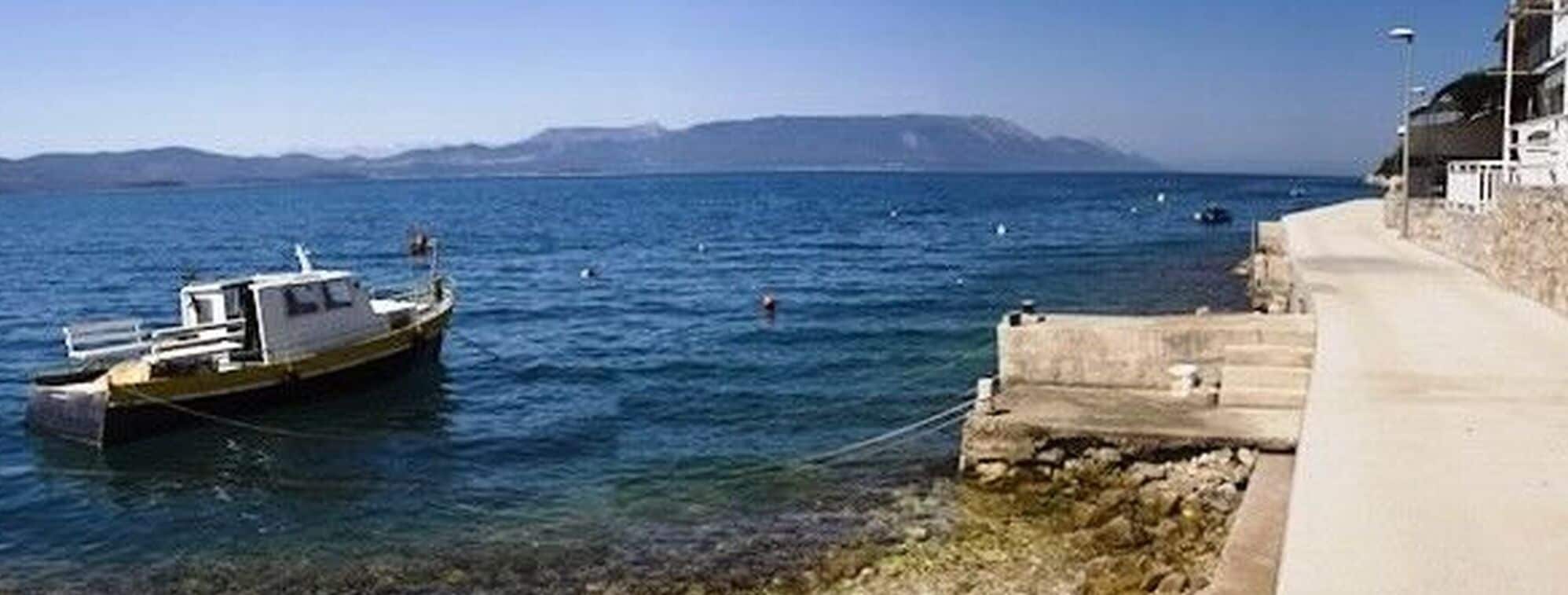 Escena costera con un bote amarrado cerca de un muelle de piedra, cielo despejado y montañas a lo lejos.