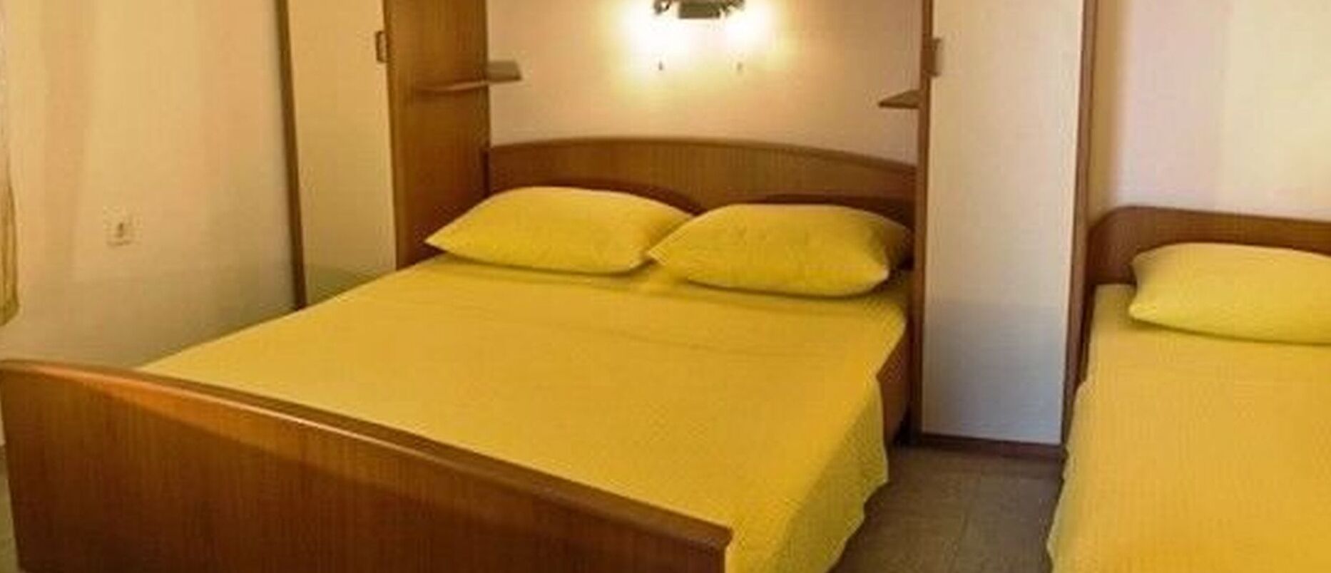 A single bed with a wooden headboard and two pillows, a wall-mounted light, and a door to the right.