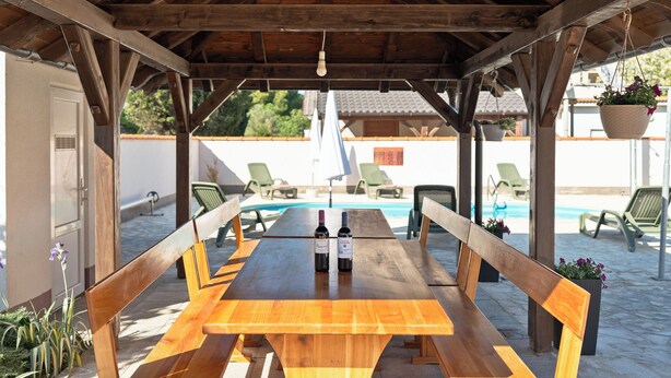 A wooden table with two bottles of wine, surrounded by benches, under a wooden roof structure. - Apartments Kralj (Privlaka)