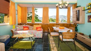 A cozy room with a sofa, dining table, and chairs, large windows with curtains, and a view of greenery and mountains. - Apartment Slavka (Kneza)