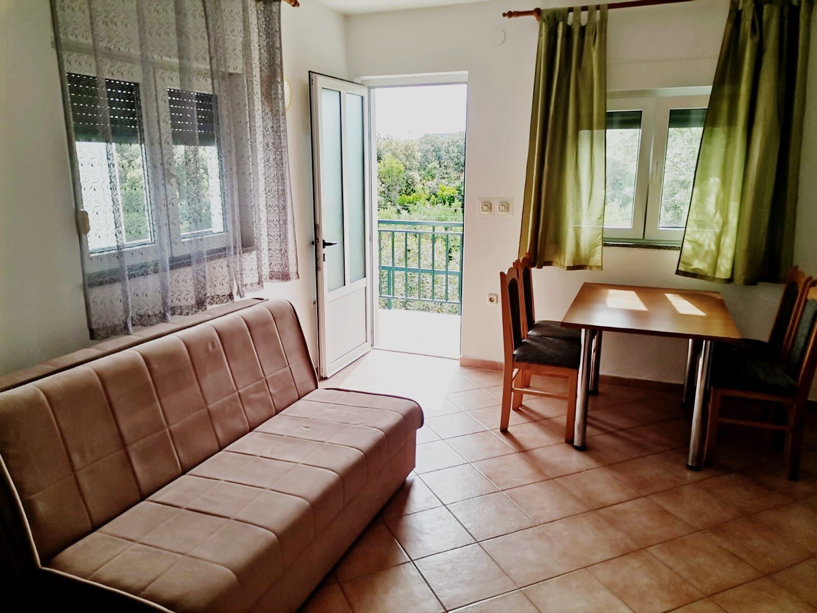 A living room with a sofa, a dining table with chairs, a sliding glass door leading to a balcony, and a view of greenery outside.