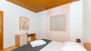 Un dormitorio con techo de madera, una cama con sábanas blancas, una mesita de noche con lámpara y una ventana con cortinas traslúcidas.