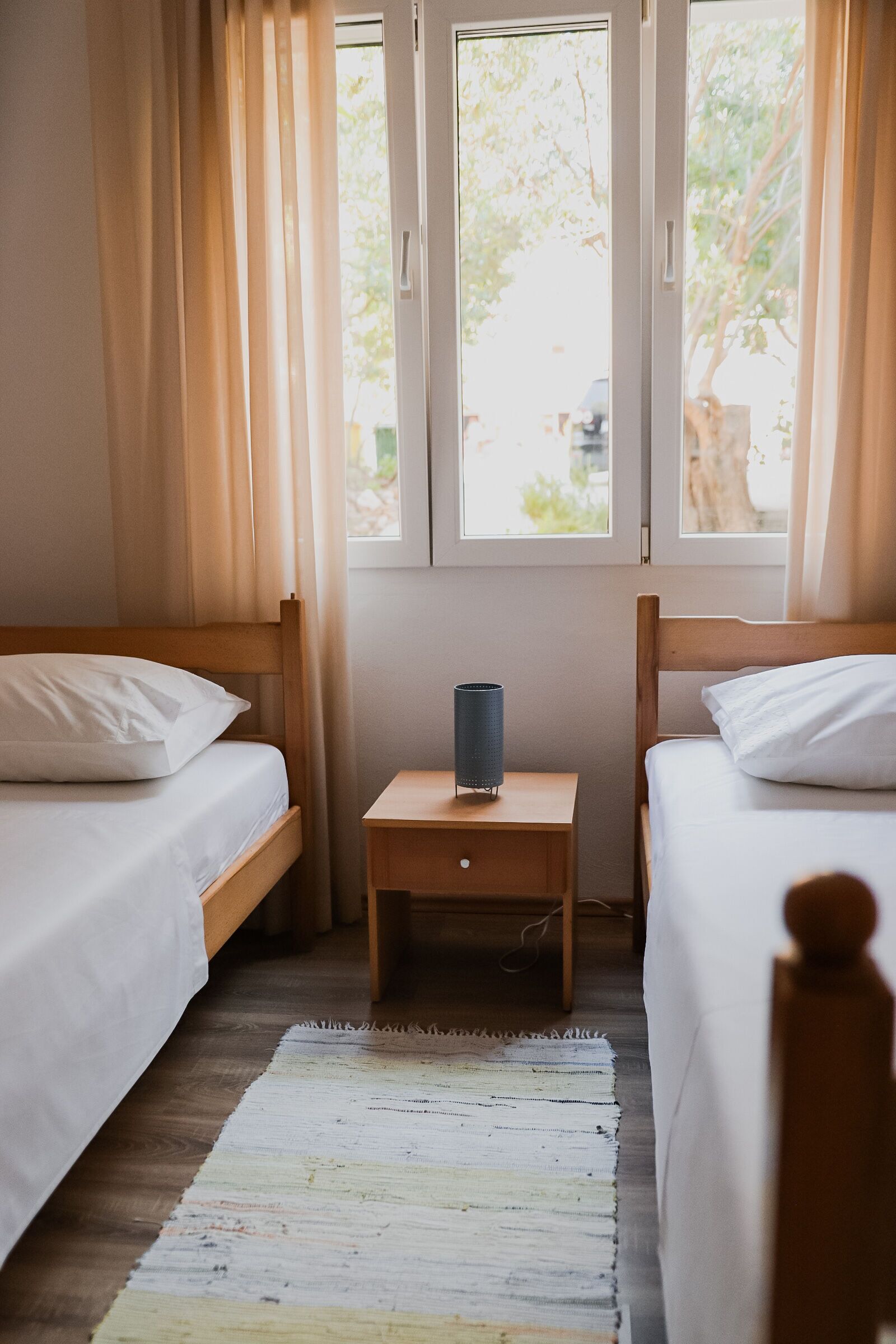 Una habitación con dos camas individuales, una mesita de noche de madera, una lámpara y una ventana con cortinas.