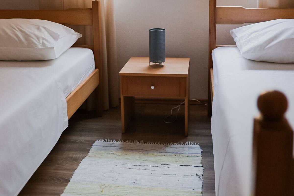A room with two single beds, a small wooden nightstand, a lamp, and a window with curtains.