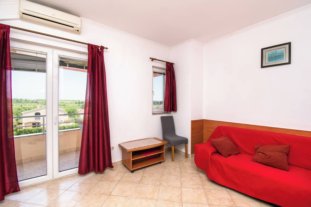 A room with a red sofa, a grey chair, a small wooden table, and a balcony with red curtains.