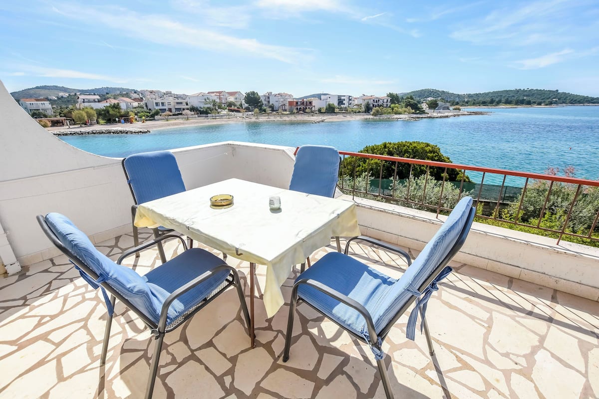 A terrace with a table and chairs, overlooking a body of water and a coastal town.