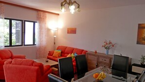 Una sala de estar con un sofá rojo, una mesa de comedor de cristal con sillas, una lámpara de pie y una ventana con cortinas.