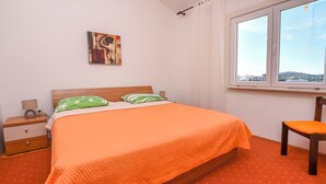 Un dormitorio con cama, una silla, una mesita de noche, un cuadro y ventana con vistas.
