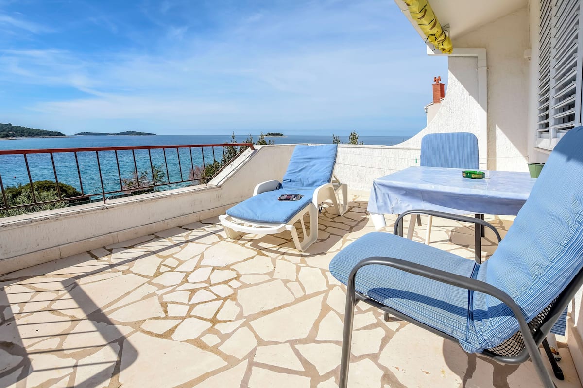 A terrace with a table and two lounge chairs, overlooking a coastal view.