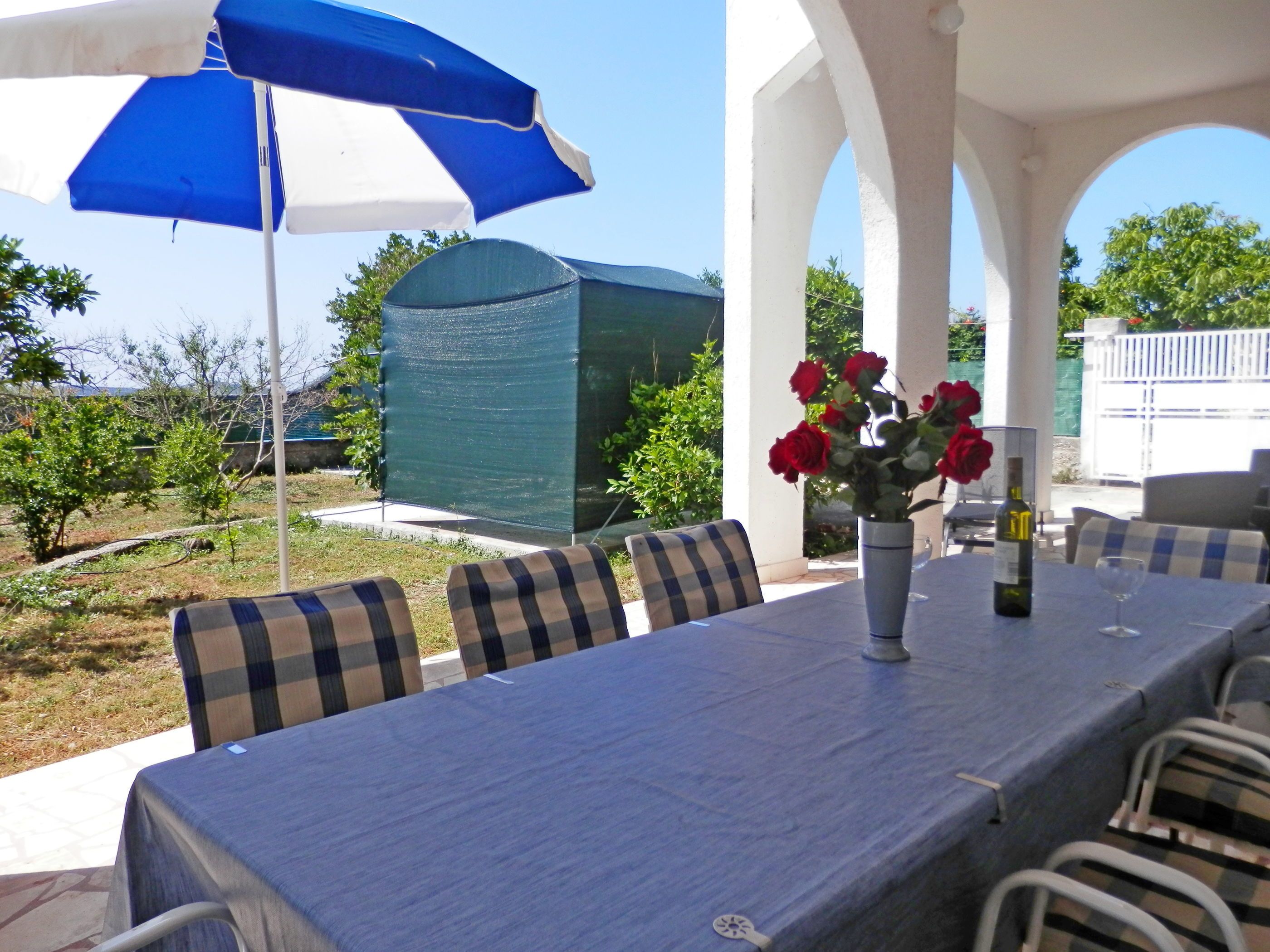 An outdoor patio with a table set for dining, covered by a blue and white umbrella, surrounded by checkered chairs, and a vase with red roses.
