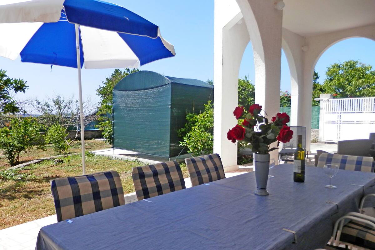 An outdoor patio with a table set for dining, covered by a blue and white umbrella, surrounded by checkered chairs, and a vase with red roses.