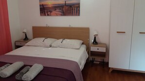 A bedroom with a bed, two bedside tables with lamps, a nightstand, a wardrobe, and a painting of a sunset over a bridge. - Apartment Dijana (Crnja Luka)