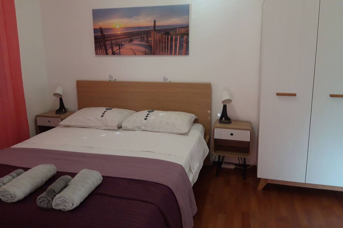 A neatly made bed with a purple bedspread, two pillows, and rolled towels on the floor. There is a painting of a sunset on the wall, a wooden headboard, and a wardrobe with two doors.