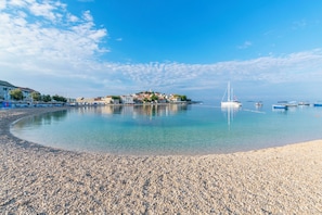 A pebbled beach with clear water, a sailboat, and buildings in the background. - Apartment Vlasta (Primosten)