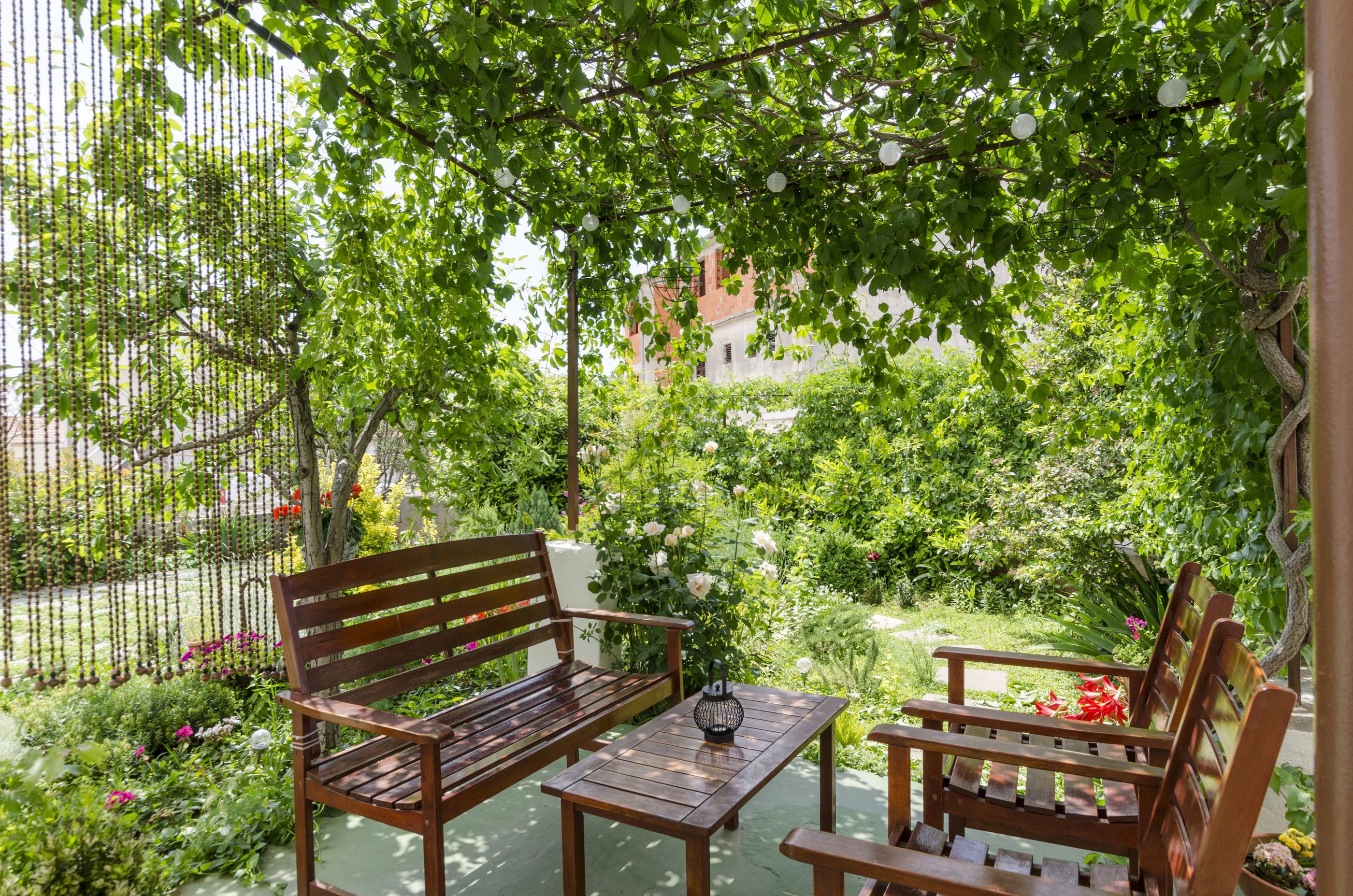Ein Holzbänkchen und Tisch in einem üppigen Garten mit einem weißen Gebäude im Hintergrund.