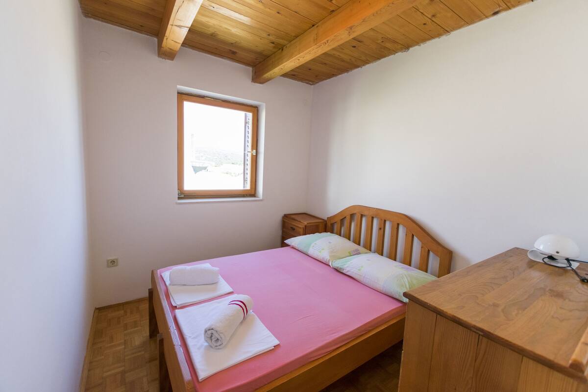 A small, clean room with a wooden bed, a bedside table, a window, and a wooden ceiling.