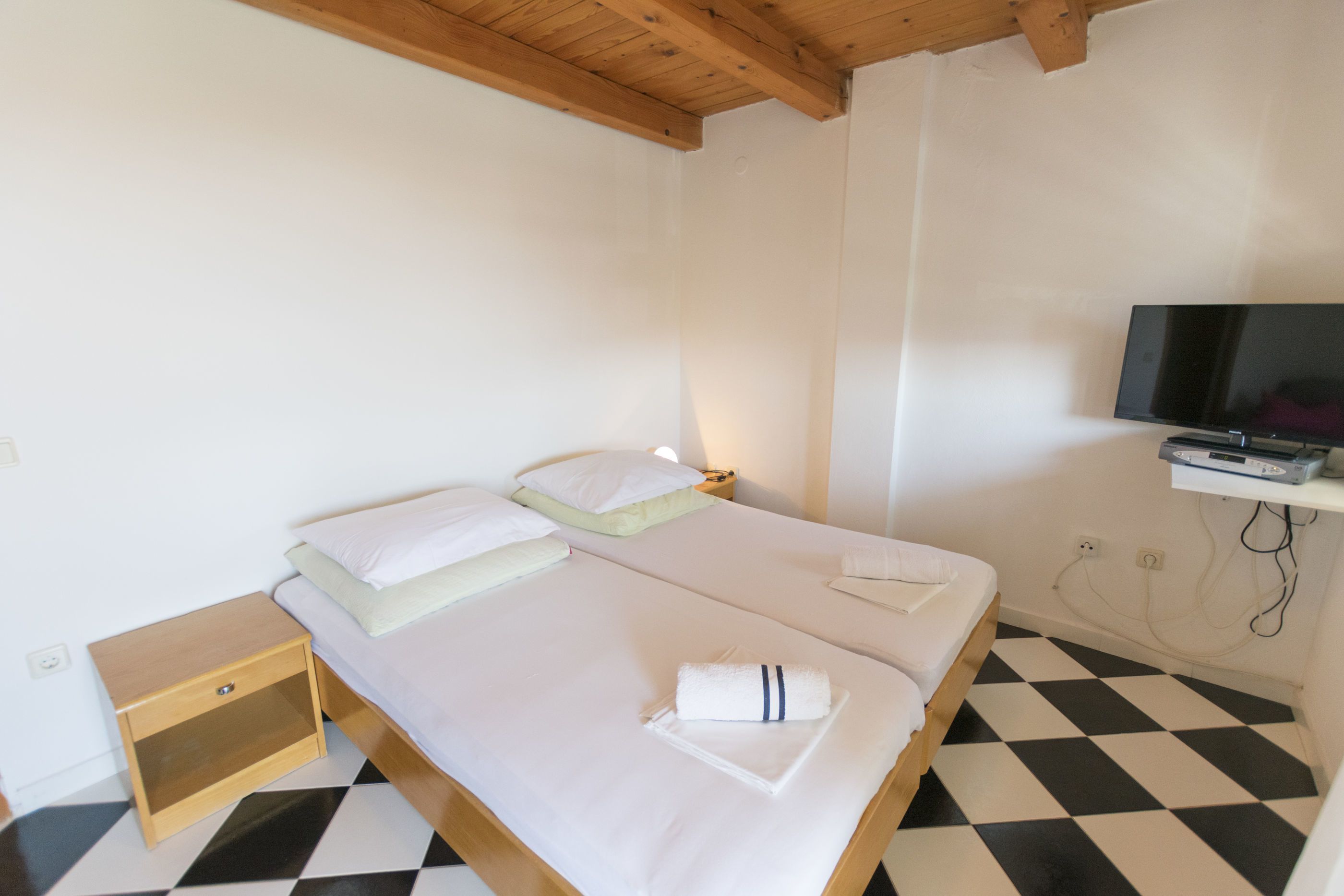 A single bed with white bedding, a wooden nightstand, a television mounted on the wall, and a black and white checkered floor.