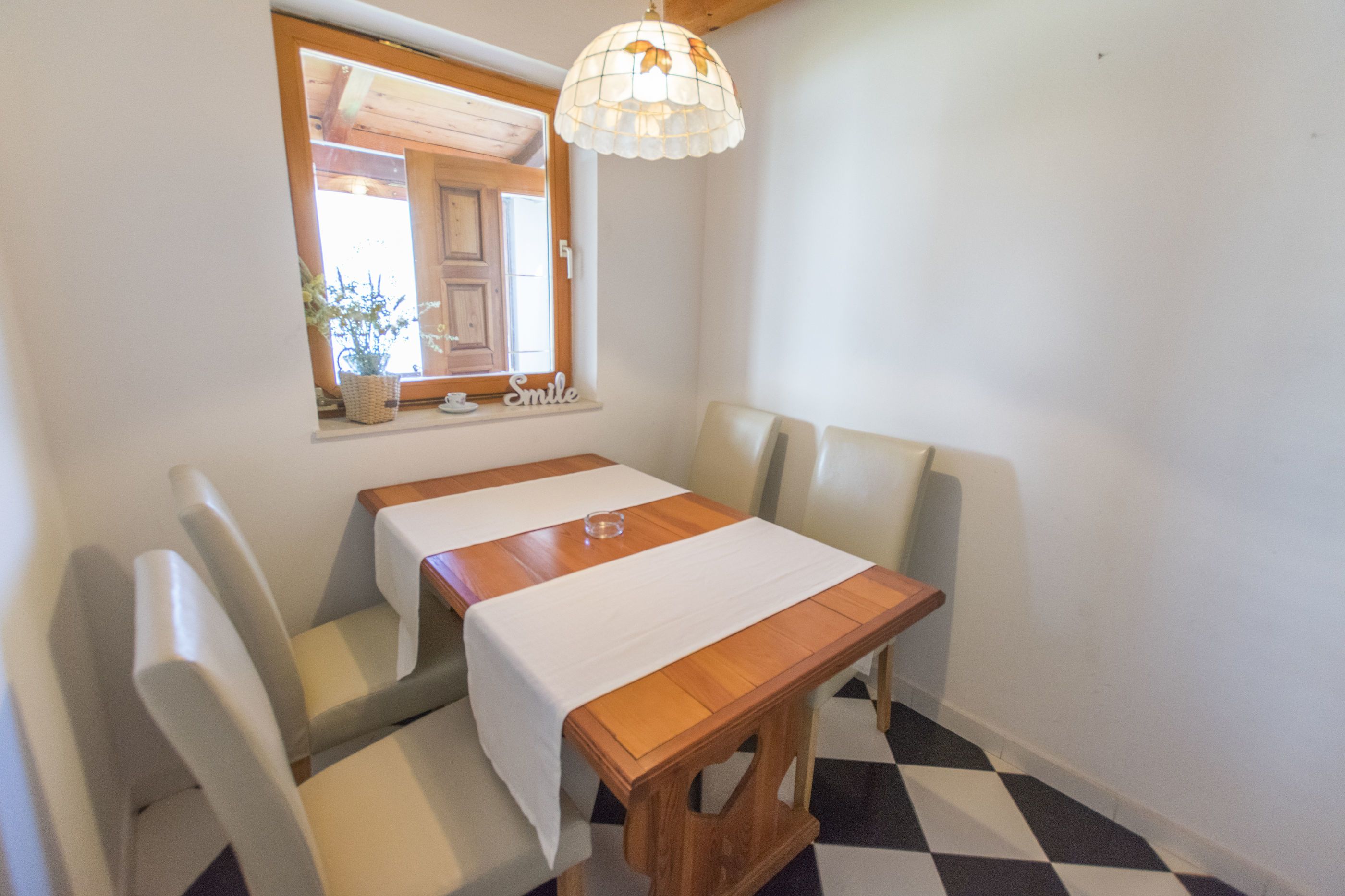 A dining area with a wooden table, white tablecloth, and four chairs. A window with a view of the outside is visible.
