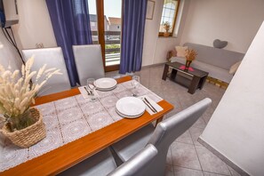 Zona pranzo con tavolo di legno apparecchiata per due persone, un vaso di fiori secchi e vista all'esterno tramite una porta vetrata.