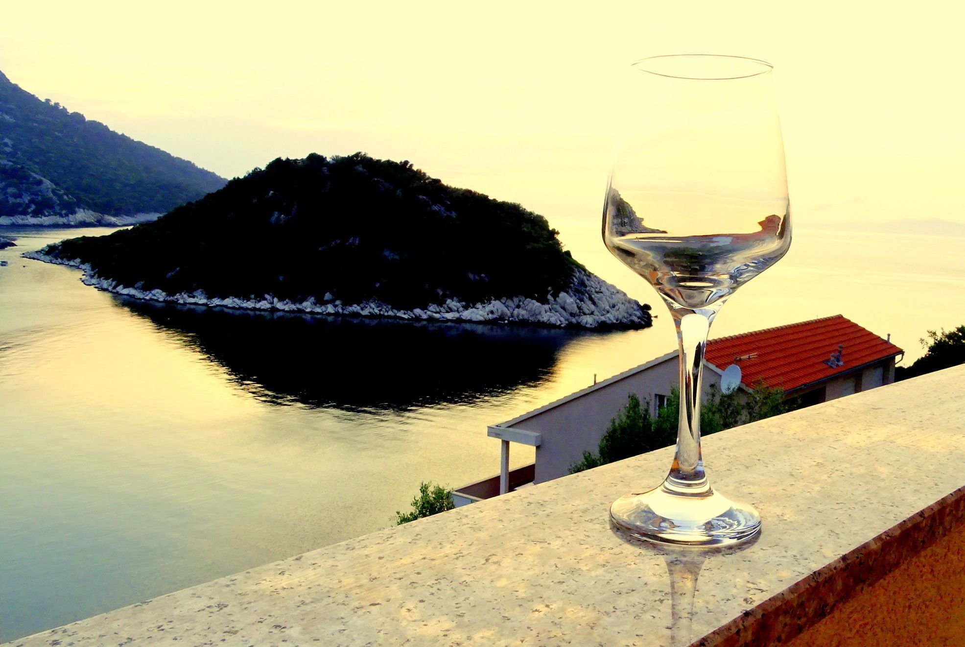 A glass of white wine on a balcony overlooking a calm lake with a small island and a building with a red roof.
