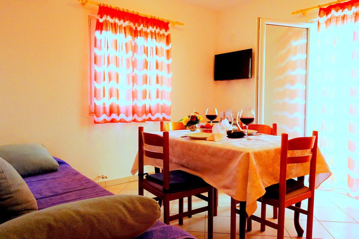 A dining area with a table set for two, a television mounted on the wall, and a window with curtains.