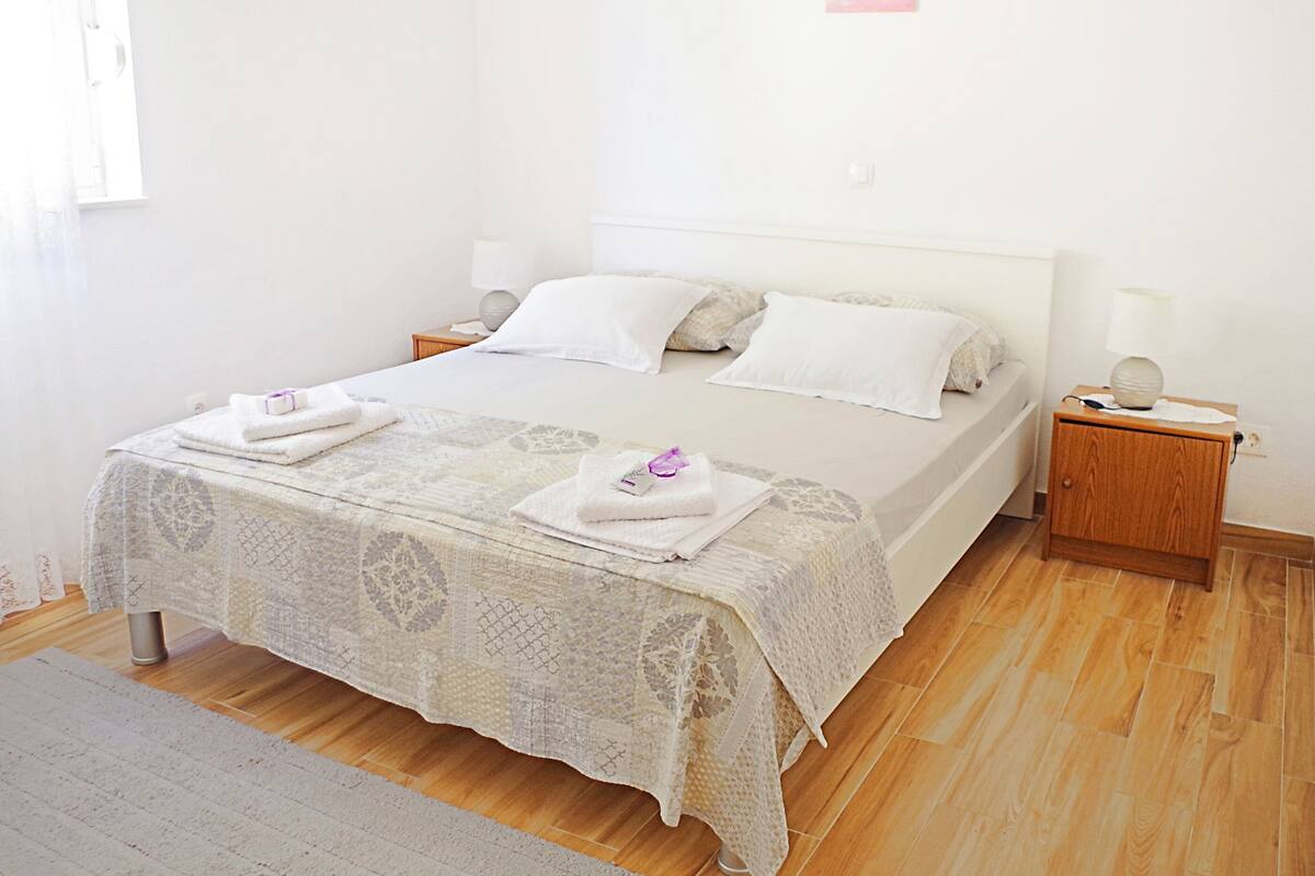 A neatly made bed with white linens, a wooden nightstand with a lamp, and a small rug on a wooden floor.