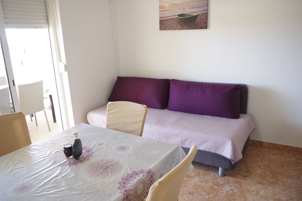 A room with a bed, a table with a tablecloth, a chair, and a painting on the wall.