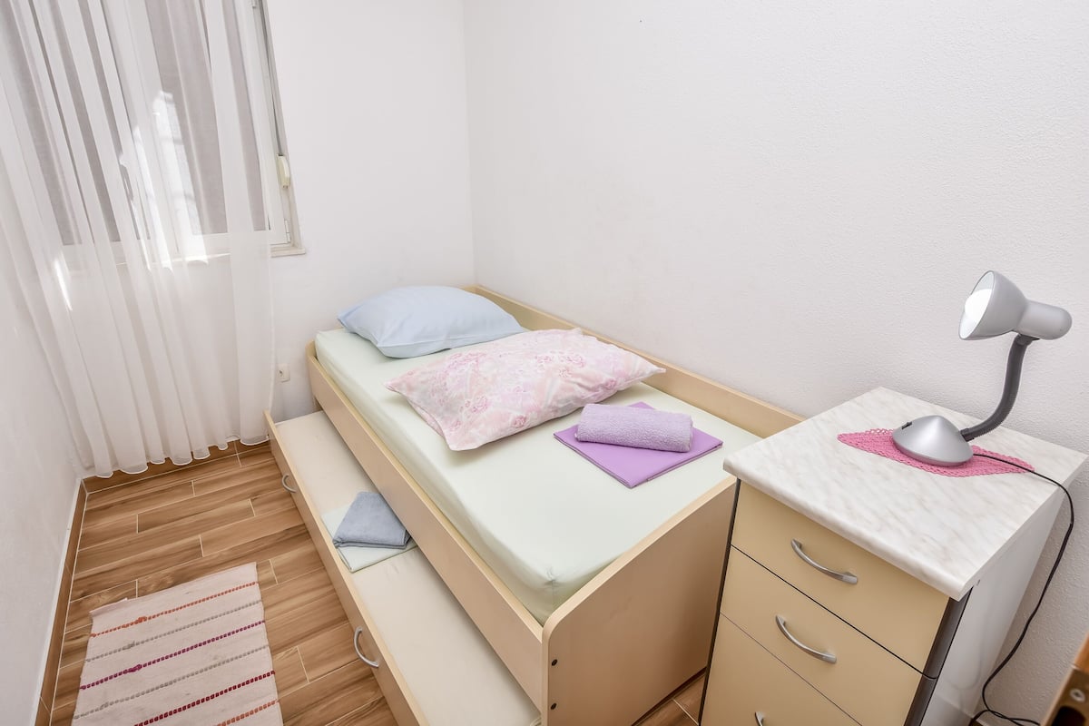 A small, neatly arranged bedroom with a single bed, a desk with a lamp, and a window with curtains.