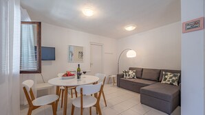 Una sala de estar moderna con un sofá gris, sillas blancas, una mesa de comedor redonda con un cuenco de fruta y copas de vino, y un televisor de pantalla plana en la pared.