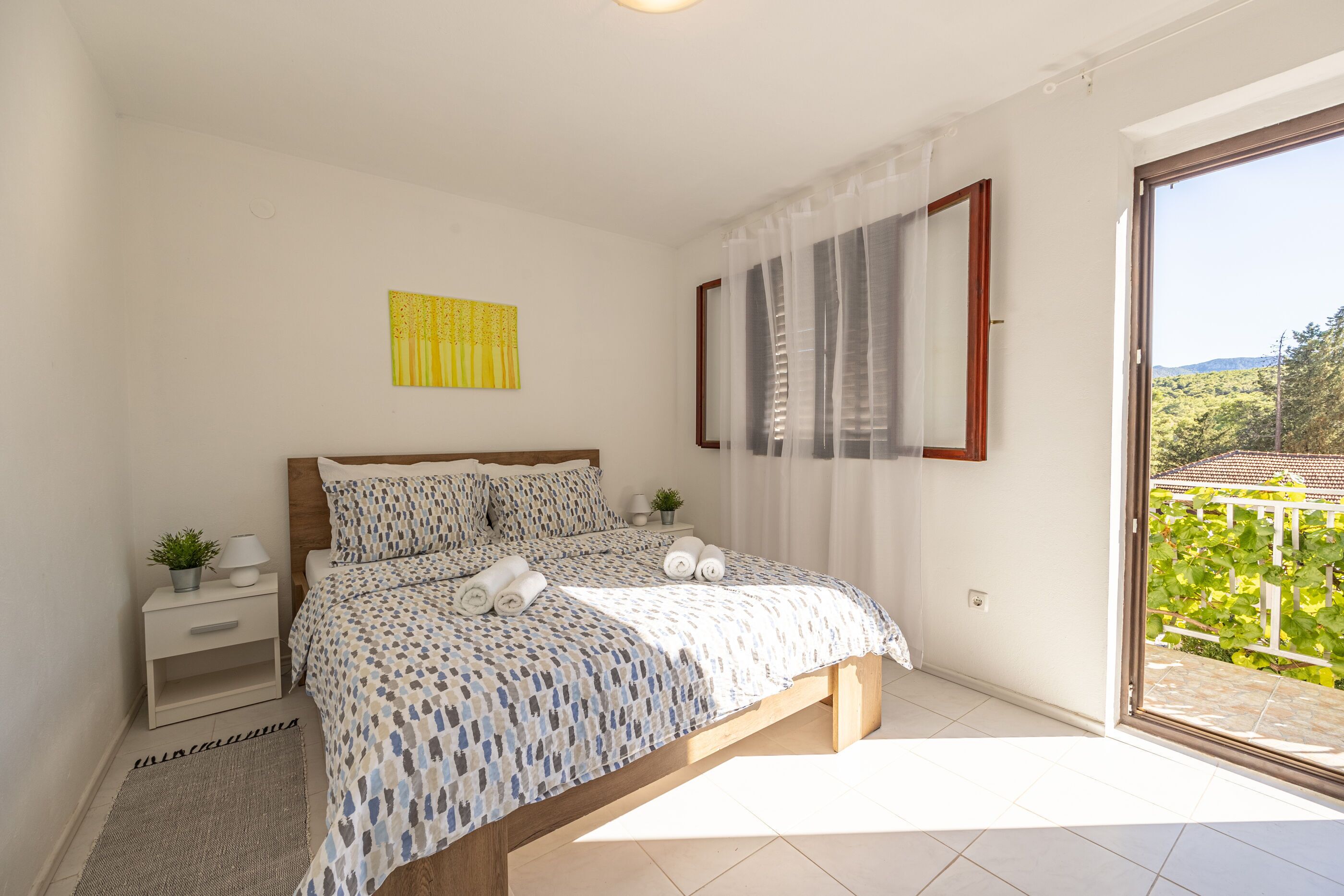 A bedroom with a bed, a nightstand, a small table with a plant, a window with curtains, and a view of greenery outside.
