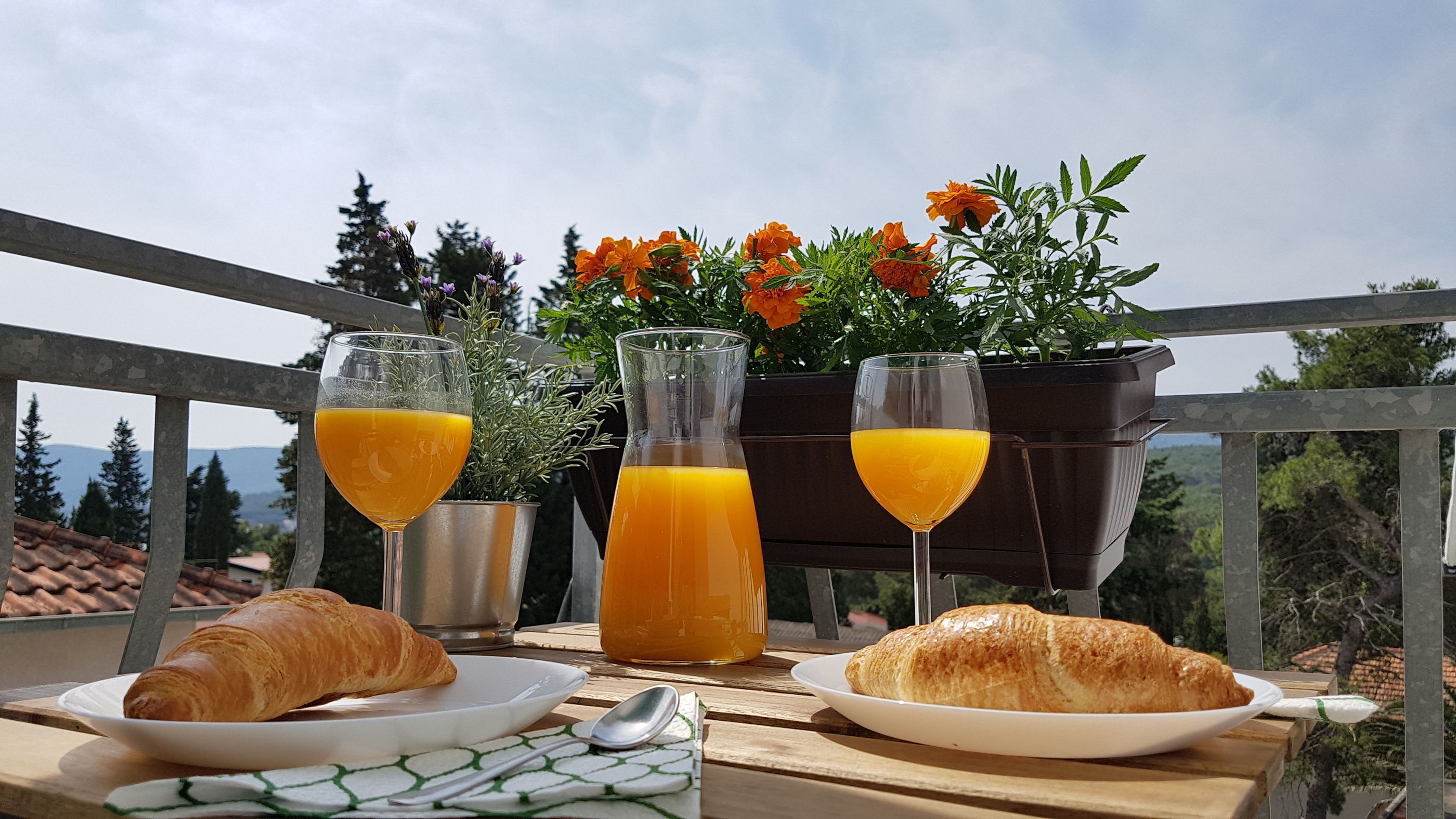 A table with croissants, two glasses of orange juice, and a bottle of orange juice on a balcony with a view of trees and mountains.
