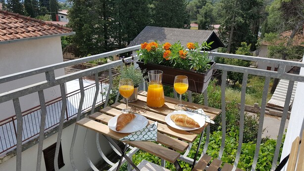 Un balcón con mesa lista para el desayuno, incluyendo cruasanes y zumo de naranja, con vistas a un jardín lleno de flores y una casa al fondo.