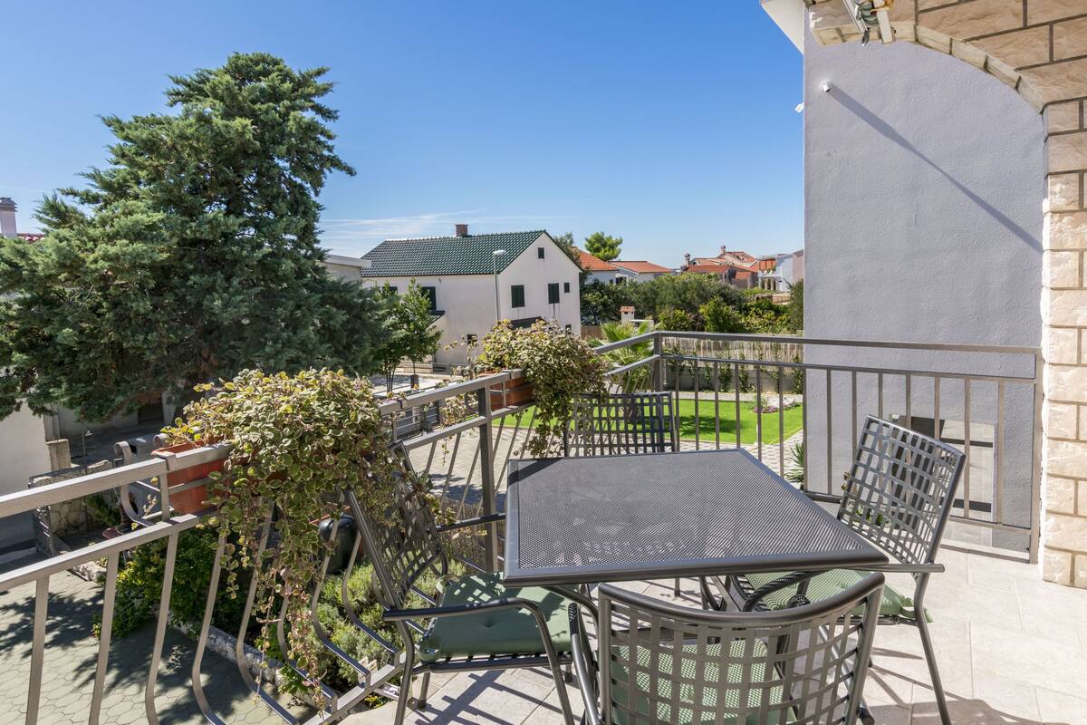 Ein Balkon mit einem Metalltisch und Stühlen, umgeben von Blumentöpfen und Blick auf Häuser.